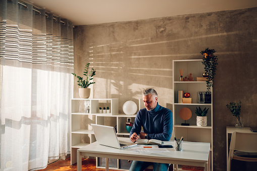 Middle aged businessman working at home while writing on agenda. Caucasian man managing home finance, reviewing bank account and using laptop. Modern home office. Copy space.