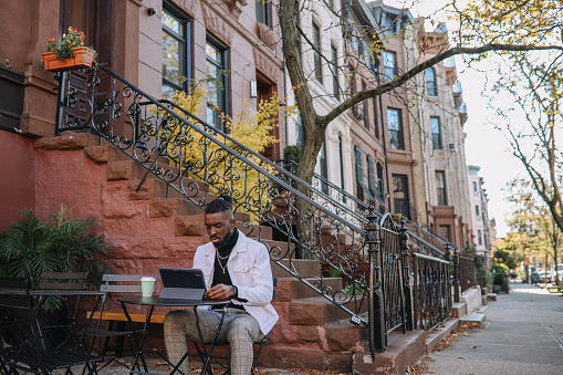 African American man sitting in a cafe in Brooklyn, NYC, having a coffee while finishing work on a digital tablet. He is doing a coding IT business, or a freelance content writing, on a sunny winter day.