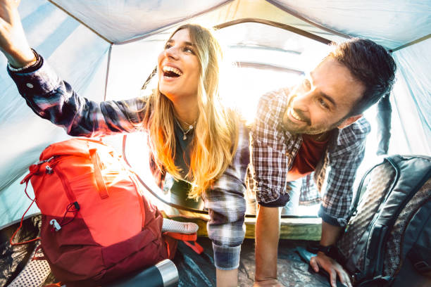 Inside pov view of happy couple at camping tent on sunset moment - Adventure and love life style concept on travel vacation - Young people having fun traveling together - Bright warm backlight filter stock photo