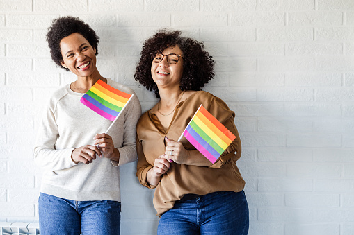 Portrait of two lesbians holding LGBTQIA flags in front of the white wall