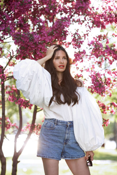 Elegant fashionable woman wearing white cotton shirt with wide sleeves lanterns flounces, high waisted denim shorts, posing near pink spring blossom trees, touches long dark hair. Outdoor portrait elegante mujer de moda con camisa blanca de algodón con volantes de mangas anchas, pantalones cortos de mezclilla de cintura alta, posando cerca de árboles de flores de primavera rosadas, toca el cabello largo y oscuro. retrato al aire libre