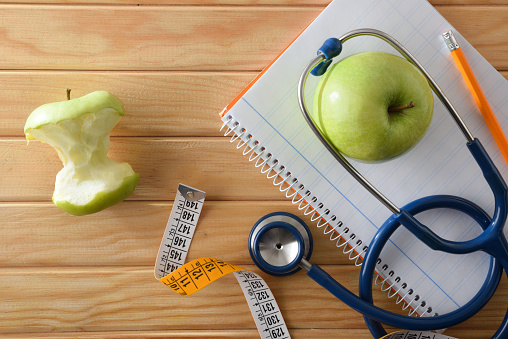 Weight control and healthy lifestyle with notebook and pencil on wooden table with apple, tape measure and stethoscope. Top view.