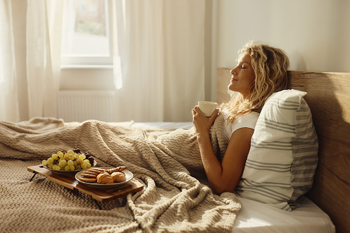 Happy woman enjoying in her morning coffee with closed eyes while relaxing on a bed. Copy space.