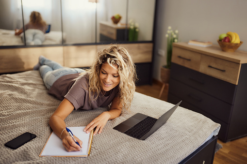 Young happy woman writing down her plans while relaxing on a bed in bedroom.