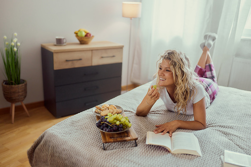 Young happy woman eating grapes while reading a book during morning time on a bed.