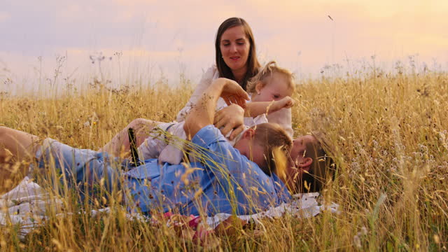 Handheld, slow motion footage of a family sitting on a picnic blanket in a meadow, father playing with kids