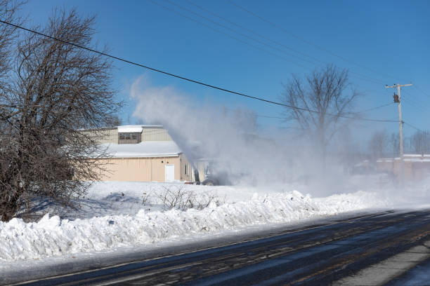 Rural road with a snow blower clearing a driveway while obscured by a cloud of fine, blowing snow. Clear sunny blue sky and bright white, fresh snow.