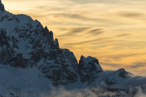 close up view of a beautiful natural environment at evening, during winter season, showing a cloudy sky, orange colored at sunset, behind a dark mountain range in silhouette, with its peaks and sides covered in snow