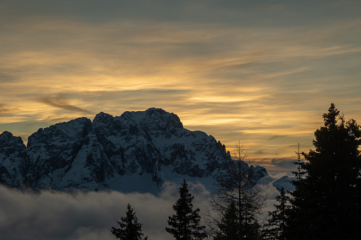 close up view of a snowy mountain range silhouette in backlight at evening. orange color and cloudy sky on the background. Val Saisera, Tarvisio (UD), F.V.G. region, Italy