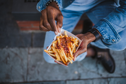 High angle shot of a man eating french fries with cheddar and bacon sitting on the street