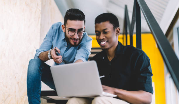istock Pensive cheerful multiethnic students using laptop on stairs in contemporary building 1460047959