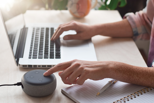 Woman sitting at desk typing on laptop computer and pressing button on virtual assistant smart speaker