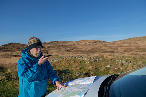 Retired man standing in front of a car while talking on a phone and looking at a map in rural Scotland