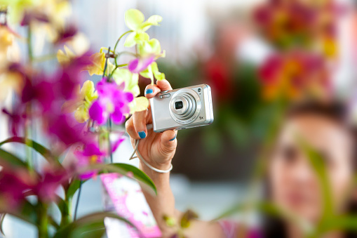 Selective focus on a photographic camera. Caucasian woman taking a picture of a flower using a grey compact camera.
