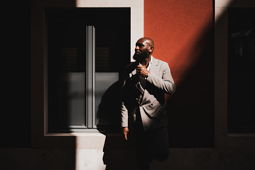 A contrast shot with a fashionable bald African American man with a neat black beard and in a grey suit stands at the junction of light and shadow in front of a reddish wall outdoors and looking aside