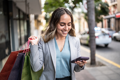 Mid adult businesswoman using phone and carrying shopping bags outdoors