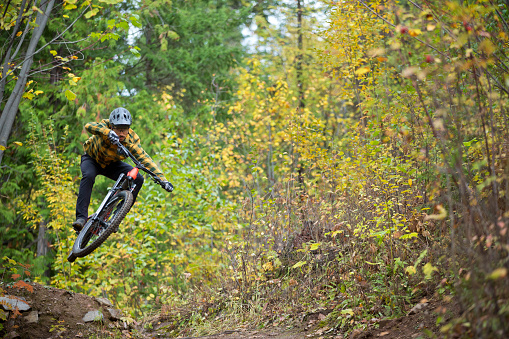 A man hits a jump during an autumn mountain bike ride in British Columbia, Canada. He is riding an enduro style mountain bike and wears a cycling helmet and casual clothing.