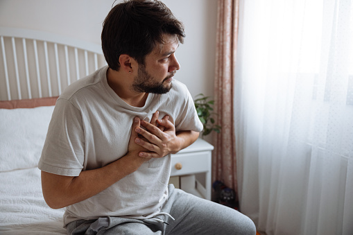 Heart attack, man with chest pain suffering at home, health problems concept. Young man with eyes closed holding his chest in discomfort, suffering from chest pain while sitting on bed at home. Elderly and health issues concept