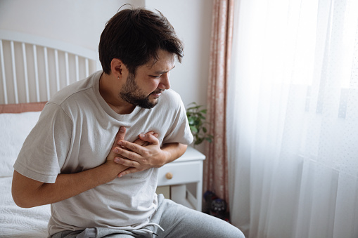 Heart attack, man with chest pain suffering at home, health problems concept. Young man with eyes closed holding his chest in discomfort, suffering from chest pain while sitting on bed at home. Elderly and health issues concept