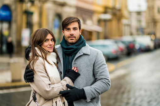 Elegant couple embracing and looking at camera while on a walk in the city on winter day.