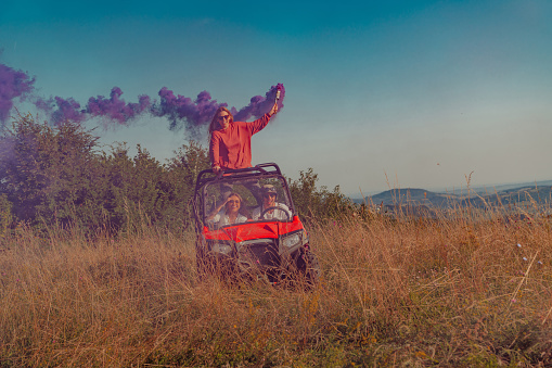 Group of young happy excited people having fun enjoying beautiful sunny day holding colorful torches while driving a off road buggy car on mountain nature