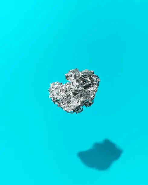 A crumpled ball of shiny silver aluminum foil floating mid-air on a turquoise blue background. Hard shadow lighting shot in a photography studio.