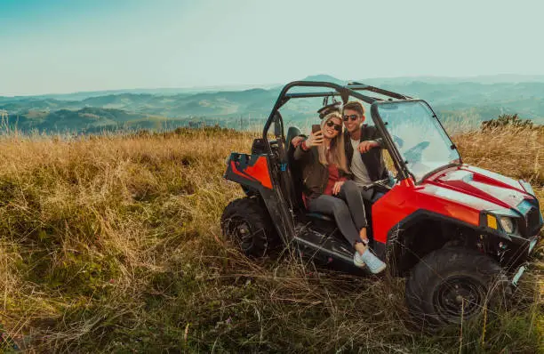 Young happy excited couple enjoying beautiful sunny day taking selfie picture while driving a off road buggy car on mountain nature.