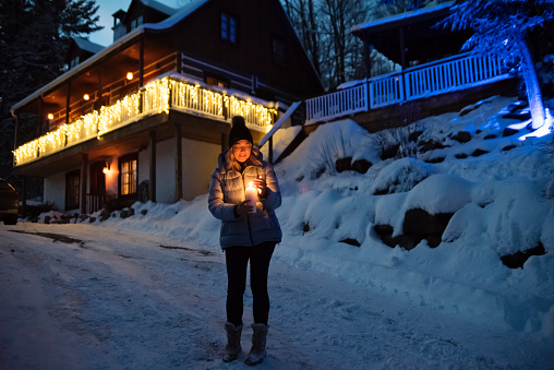 Mature woman holding a candle outdoors at a chalet in winter night. She is in her fifties, and is wearing a pale blue winter coat and a black wool hat. Horizontal full length outdoors shot with copy space. This was taken in the north of Quebec, Canada.