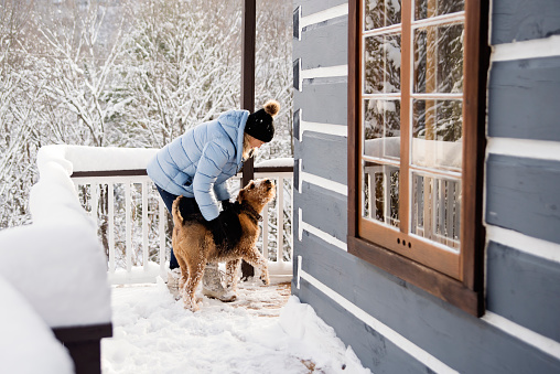 Mature woman greeting her dog on the chalet balcony in winter. She is in her fifties, and is wearing a pale blue winter coat and a black wool hat. Dog is an Airedale. Horizontal full length outdoors shot with copy space. This was taken in the north of Quebec, Canada.