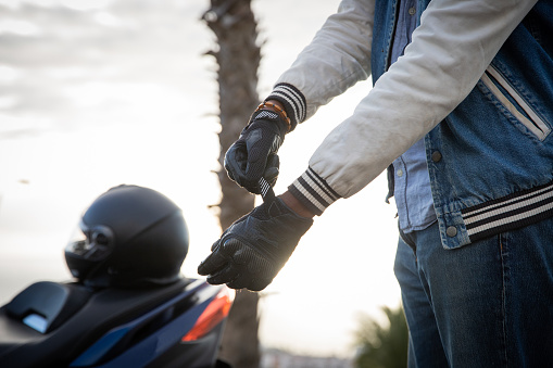 Hand of a biker putting on gloves with the scooter parked to the side with helmet on it