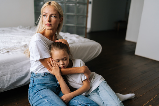 Portrait of upset cute little girl and loving caring mother looking away comforting offended afraid child daughter, showing love and care, expressing support, hugging, stroking hair sitting on floor.