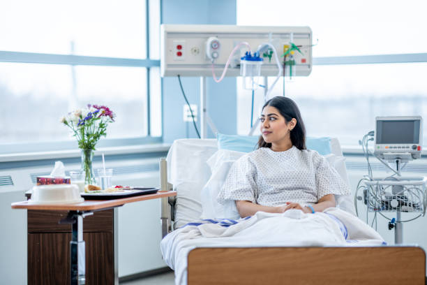 Woman in the Hospital A young woman of Middle Eastern decent sits up in her hospital bed after surgery. She is wearing a gown and is tucked warmly under a blanket and she looks down contemplatively. sick woman hospital bed stock pictures, royalty-free photos & images