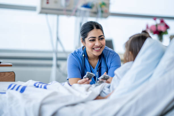 Nurse Checking on a Patient A female nurse of Middle Eastern decent sits at the edge of a hospital bed as she check in on her young patient. She is wearing blue scrubs and is attempting to cheer the young girl up. body care stock pictures, royalty-free photos & images