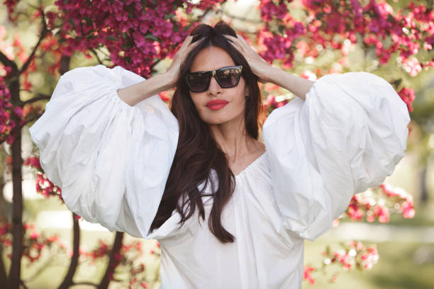 hermosa mujer morena hermosa con cabello largo en camisa blanca de algodón con linternas de mangas anchas vuela sobre fondo de flores de sakura de primavera. modelo toca el cabello - puffed sleeve fotografías e imágenes de stock