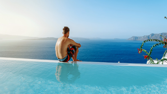 Santorini Greece Oia, young men in swim shorts relaxing in the pool looking out over the caldera of Santorini Island Greece, infinity pool, a young guy on a luxury vacation in Europe Greece