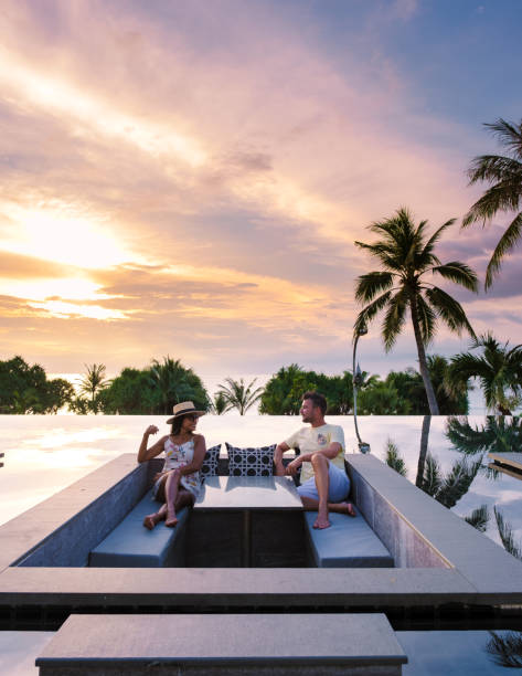 couple watching sunset in infinity pool on a luxury vacation in Thailand couple watching the sunset in an infinity pool on a luxury vacation in Thailand, man and woman watching the sunset on the edge of a pool in Thailand on vacation travel hotel accommodations stock pictures, royalty-free photos & images
