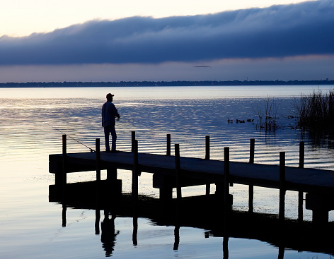 A Single Male Fisherman Stands on the Freshwater Lake Dock Fishing in the Pre-dawn Light