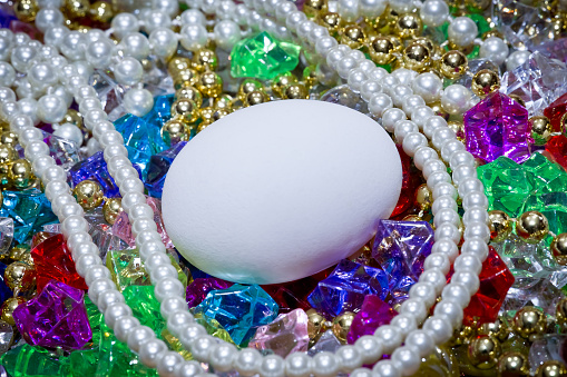 Close-up photo photo of a white chicken egg on top of multi-colored jewels, pearls, diamonds, crystals, gold beads and jewelry. Copy space.