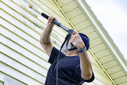 Real life, real person DIY senior man colorectal cancer survivor is back to normal routines after a few years of medical problems. He is cleaning the clapboard siding on his house with high pressure cleaning power wash equipment. The dark blue symbolic colorectal cancer support wristband labeled 
