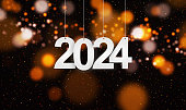 Happy New Year 2024 with small glitters sprinkling down. Hanging white paper cut number with festive confetti on an orange golden blurry bokeh background.