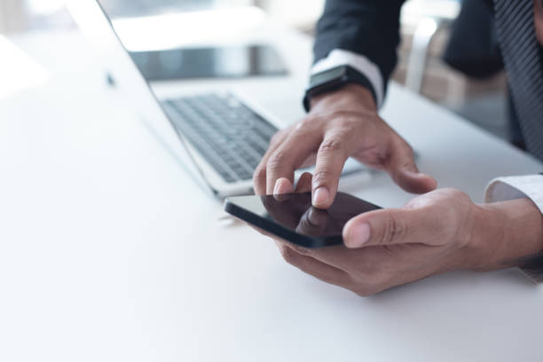 Businessman in black suit using mobile smart phone and during working on laptop computer at office. Business man finger touching on smartphone screen with laptop on office desk, close up hombre de negocios con traje negro usando un teléfono inteligente móvil y durante el trabajo en la computadora portátil en la oficina. hombre de negocios tocando con el dedo en la pantalla del teléfono inteligente con la computadora portátil en el esc