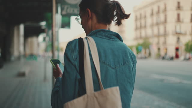Young woman with ponytail wearing denim jacket with mobile phone in her hands walks along the streets in cityscape background. Girl browsing information on smartphone