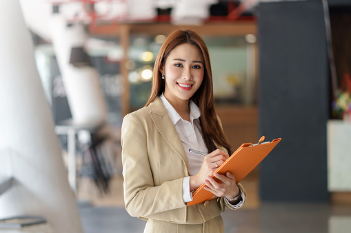 Charming young Asian businesswoman with a smile standing holding document clipboard at the office. Looking at camera.