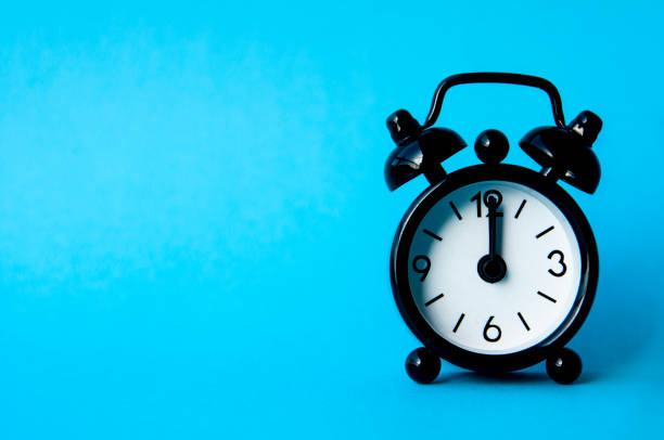 istock Black alarm clock isolated on blue background. The clock set at 12pm. Copy space and time concept 1460175528