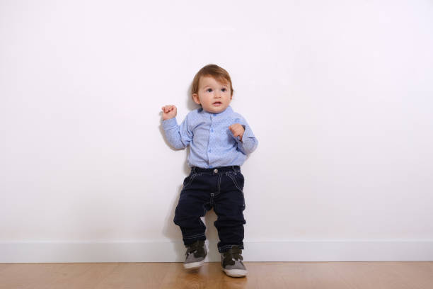 Portrait of adorable curly baby toddler boy with brown eyes dressed in beige pants, sweater and sneakers. Child sits against a white background and looks away with a smile.