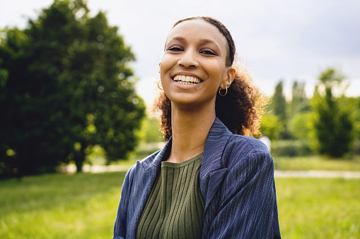 Genuine outdoor portrait of an hipster African American woman with nose and lip piercing and curly hair tied into a ponytail standing carefree in the park - individuality people lifestyle concept