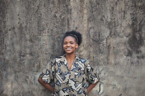 A portrait of a young smiling black woman with short curly hair leaning on a wall outdoors