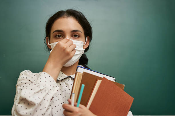 istock Multi-racial female teacher fixing her facemask and holding her textbooks in a classroom - covid19 1460189526