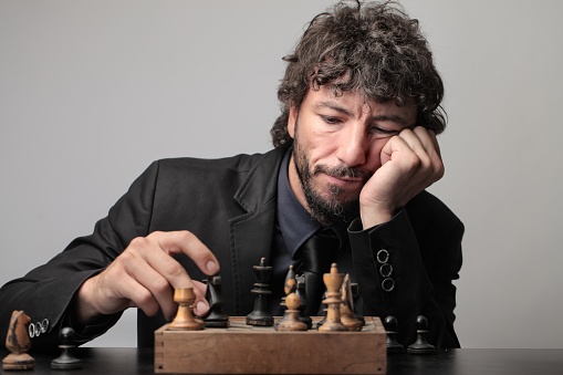 An adult Italian man with curly hair wearing a suit and playing chess with himself