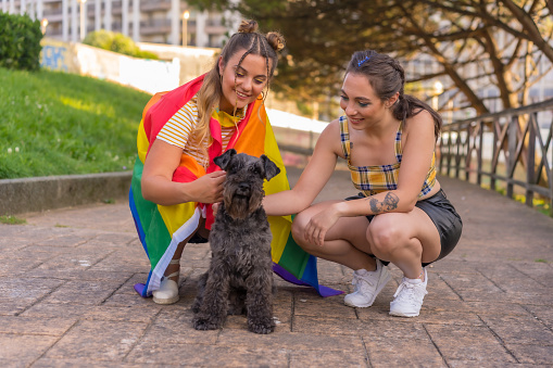 A closeup shot of two young lesbian females with a rainbow flag petting a cute dog
